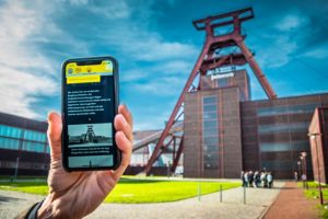 Eine Hand hält ein Smartphone mit der App Perspektivwechsel vor der Zeche Zollverein in die Höhe.